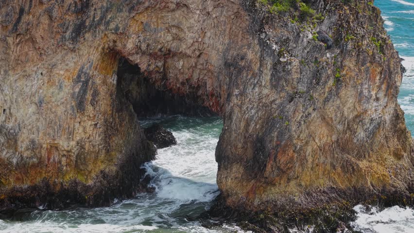 Aerial drone view of a grass-covered sea stack with a sea cave on Chiloé Island, Patagonia’s Pacific coast, Chile. Powerful waves hit a remote sandy beach and rugged green cliffs under an overcast sky