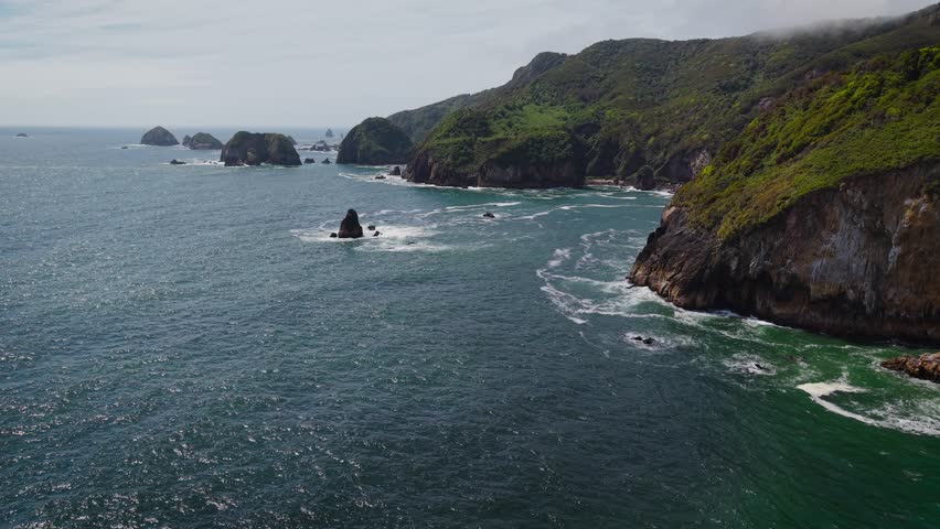 Aerial drone view of a grass-covered sea stack with a sea cave on Chiloé Island, Patagonia’s Pacific coast, Chile. Powerful waves hit a remote sandy beach and rugged green cliffs under an overcast sky