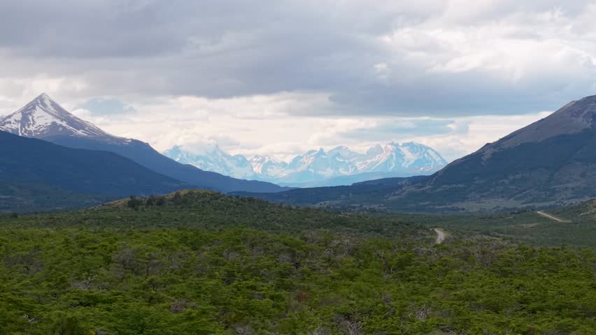 Distant view of Torres del Paine peaks in Chilean Patagonia, framed by dark mountains and green scrubland with a winding road under dramatic clouds.