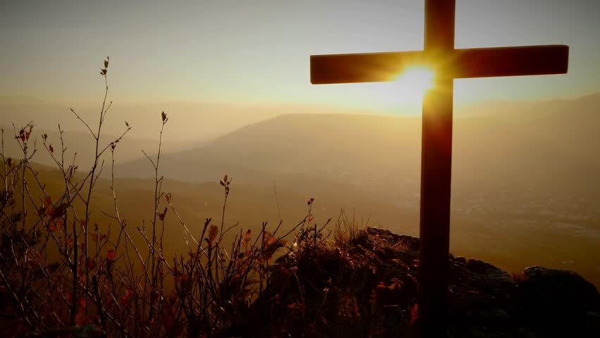 Wooden Christian Cross atop rocky hill at sunset. Sun shines through, bathing landscape in golden light. Mountains in background, representing faith, hope, and Easter.
