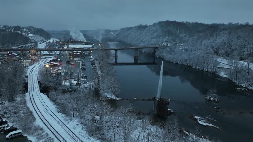 Snowy aerial view of river and fountain in Lynchburg, showing winter landscape with bridge, calm water, snow-covered hills, rail lines and moody blue dusk atmosphere. Wide shot.