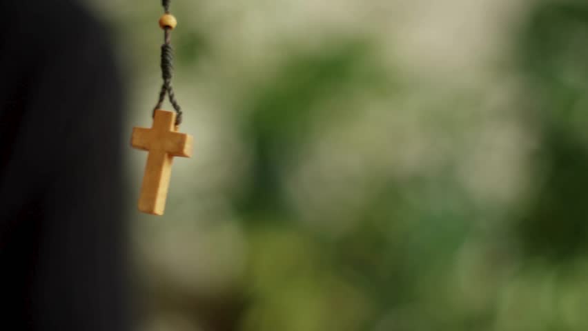 Wooden cross pendant dangles gently in natural light. Blurry green foliage visible behind it. Peaceful, spiritual imagery. Suitable for religious themes and reflective content.