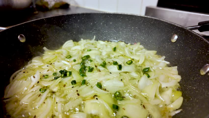 Sliced onions and green chillies frying in hot oil, creating steam in a pan