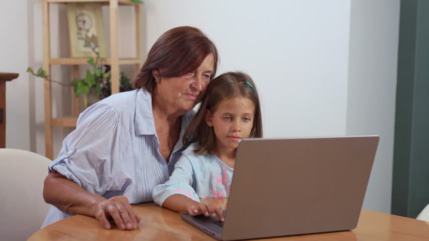 Grandmother and granddaughter sitting at table at home using laptop for elearning, sharing a focused and interactive moment while exploring digital education together