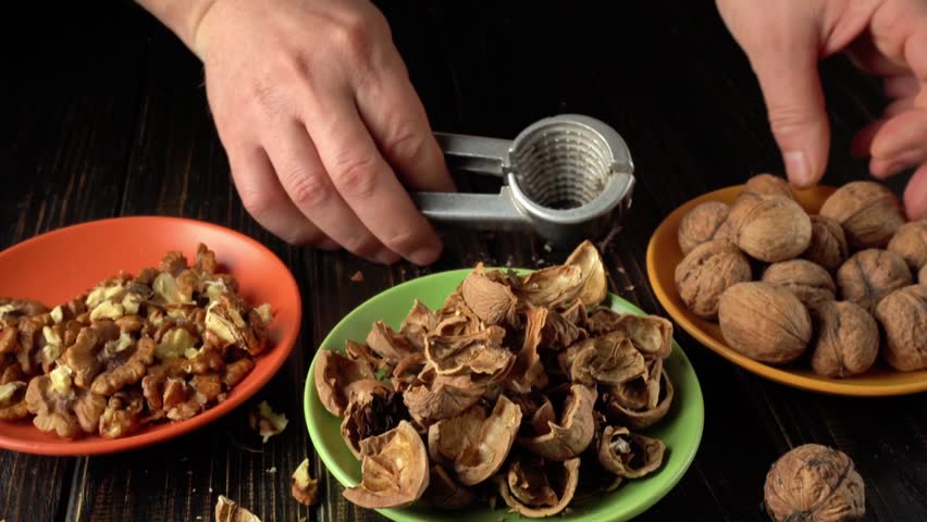 Hands using a nutcracker to crack open walnuts, showing the nut inside while empty shells and whole walnuts are arranged on colorful plates nearby