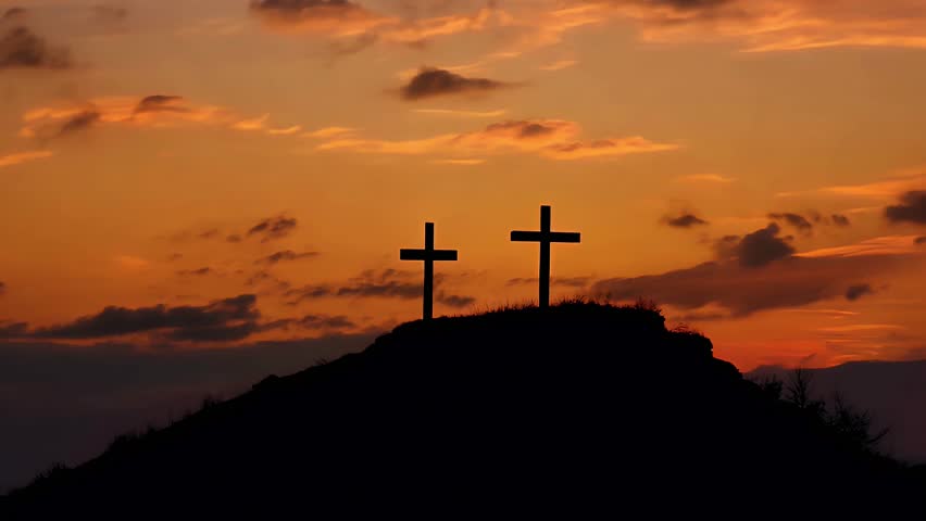 Two silhouetted crosses stand atop a hill as golden sunset paints sky with beautiful orange hues and dramatic clouds. Peaceful and spiritual.