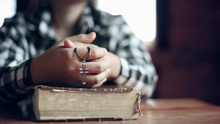 Close up shot. Person wearing a cross necklace, holding clasped hands over an old, open Holy Bible. Prayer and faith concept. Wooden table background.