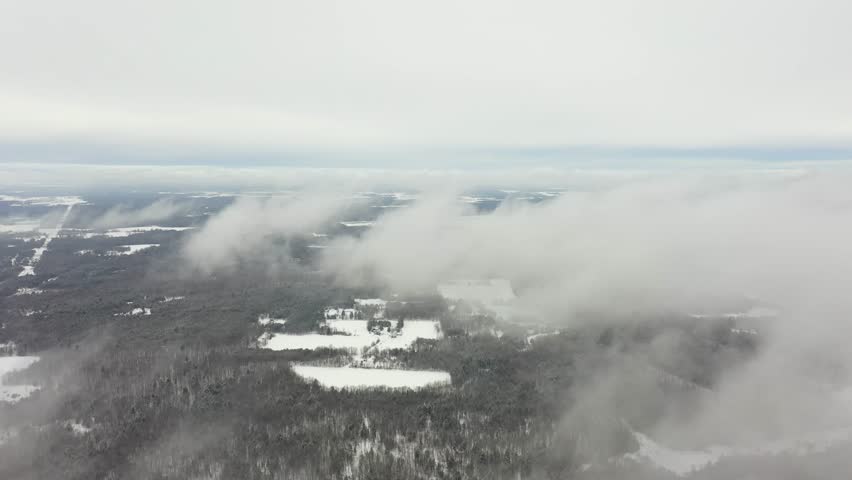 Stunning aerial perspective flying through low, wispy clouds over a vast, snow covered forest and frozen lakes, revealing a serene and cold winter landscape on an overcast day