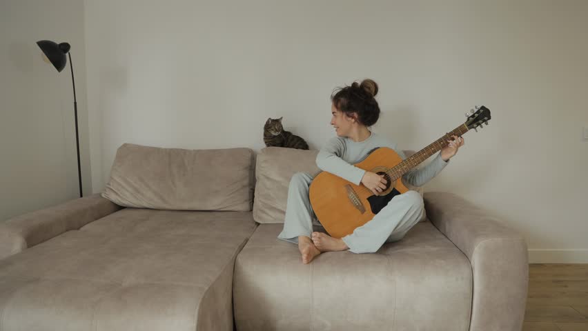 A young woman sits on a couch with her cat and plays a guitar, enjoying her music.