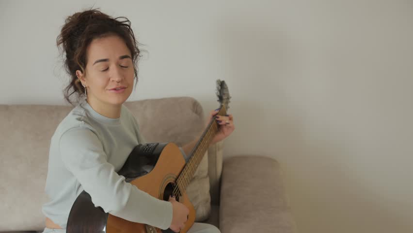 A young woman sits on a sofa and plays an acoustic guitar. She smiles at the camera and seems to be enjoying herself.