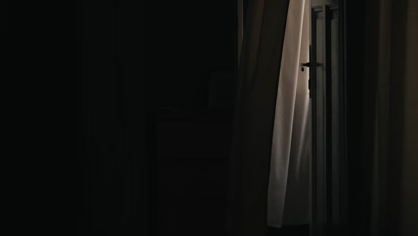 In the dark bedroom, the window is open and the curtains flutter. A calm, quiet stillness reigns in the room. The curtains flutter gently in the wind.
