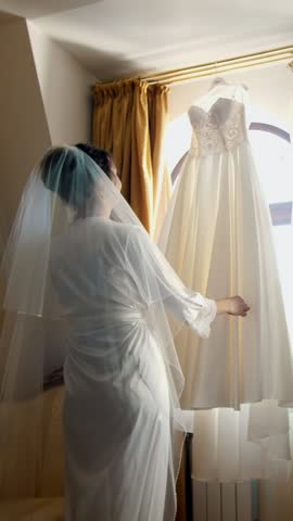 near window, in room, white wedding dress hanging on the window eaves. the bride, a beautiful girl in a white peignoir, a bathrobe, is considering her wedding dress, preparing for the wedding