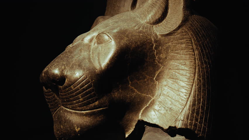 Close-up of an ancient Egyptian stone sculpture, a tiger-like head with dramatic lighting, showing carved details, weathered surface, and timeless craftsmanship against a dark background.