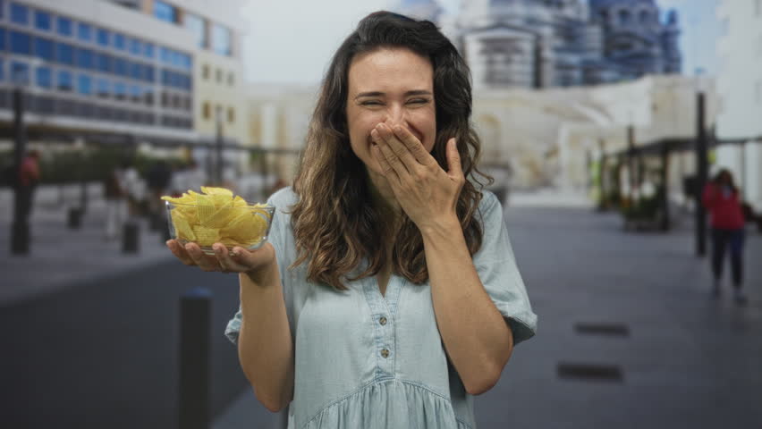Woman smiling and holding glass bowl of crispy chips with hand to mouth gesture on street; happiness.