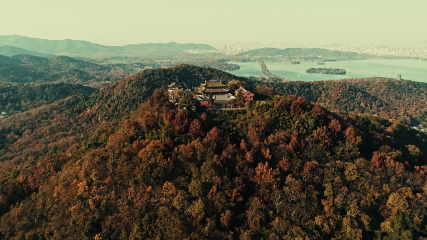 Hangzhou, China. Aerial view of autumnal landscape with temple amidst forested hills and serene lake, with cityscape in background.