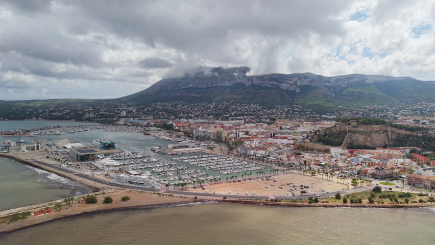 Aerial panoramic view of Denia marina and coastal cityscape with Mediterranean Sea, sandy beach, harbor, and dramatic clouds over mountain backdrop. High quality 4k footage
