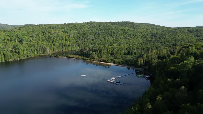 Aerial glide over quiet forest lake with smooth water surface and uninterrupted tree line, calm dock with cloud reflection