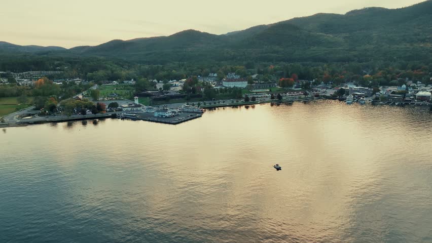 Adirondack Mountain, New York State. Aerial view of a serene lakeside town during sunset, with mountains in the background. The town is nestled amidst a forested hillside.