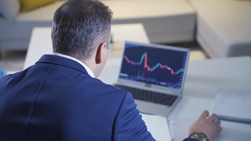 A businessman is focused on analyzing cryptocurrency market trends displayed on his laptop screen. The setting is a modern office environment.
