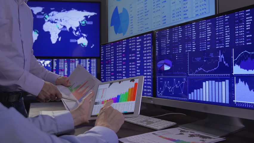 Two business professionals are reviewing financial charts and graphs displayed on multiple monitors in a modern office setting. The screens show various data related to forex and cryptocurrency markets.