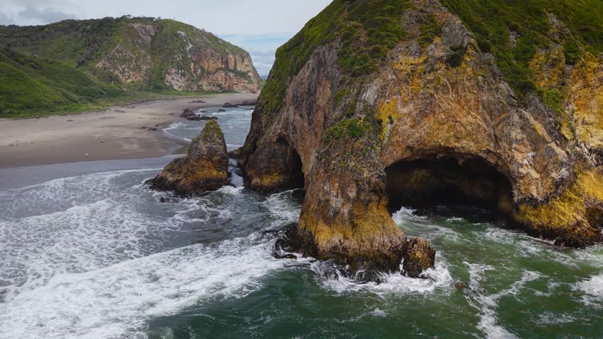 Aerial drone view of a grass-covered sea stack with a sea cave on Chiloé Island, Patagonia’s Pacific coast, Chile. Powerful waves hit a remote sandy beach and rugged green cliffs under an overcast sky