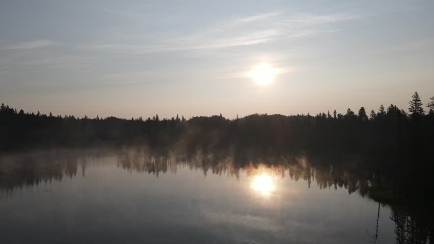 Drone flying low over a calm northern lake at sunrise with the sun reflecting in the water
