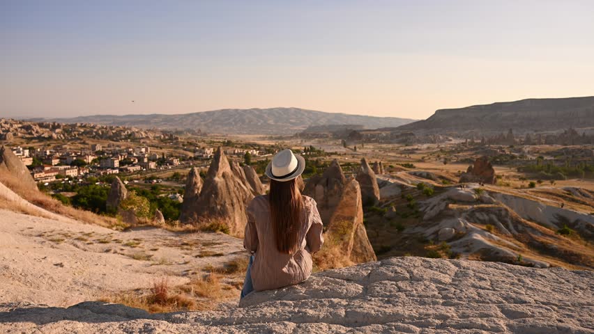 Hiker in hat enjoys picturesque views of valley in Cappadocia. Travel blogger sits on rocky ledge and enjoys panoramic sunset view in Goreme. Travel concept, UNESCO World Heritage Sites in Turkey. 