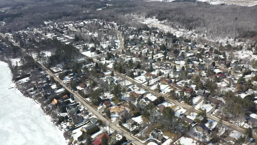 Expansive aerial perspective showing a quiet suburban community with houses and streets covered in snow, situated along the shoreline of a vast, frozen lake on a bright winter day