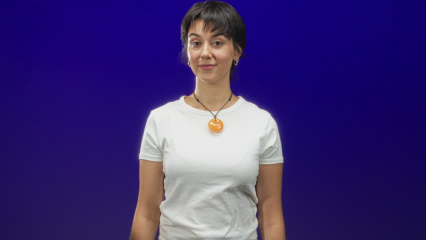Woman with short hair and orange pendant points finger to nose while smiling, wearing white t shirt in studio; playful curiosity.