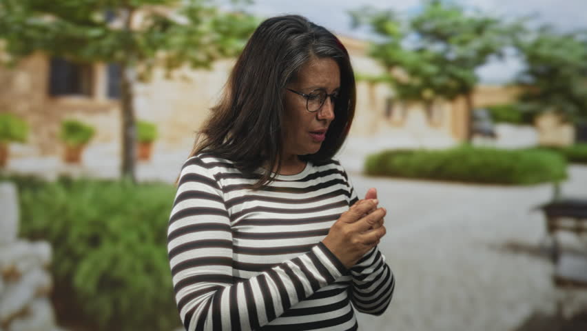 Woman clasping hands and touching her face in front of building, wearing striped top and glasses; reflection concern.