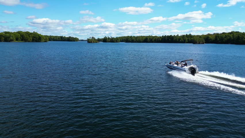 Wide aerial orbit of small boat moving steadily across blue lake with forested shoreline, Canada landscape