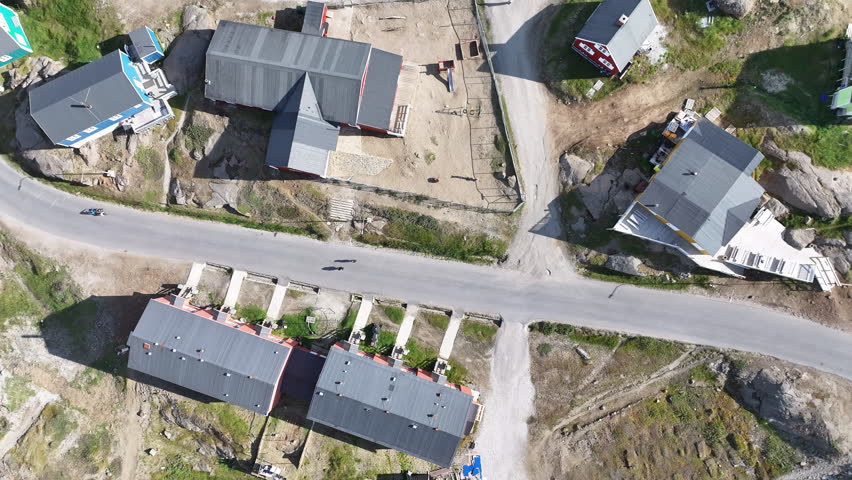 Drone footage looking down at the streets and colorful wooden houses of Tasiilaq, Greenland. The shot reveals the rocky terrain, winding roads, and residential architecture of the remote Arctic settlement during a sunny summer day.