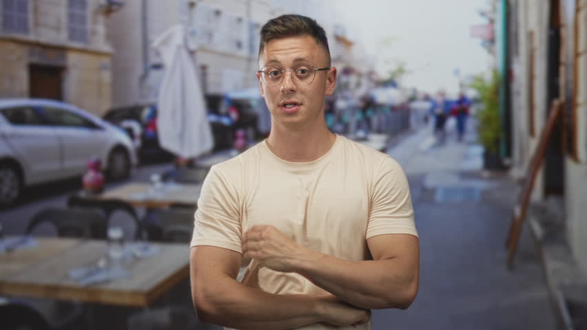 Young man with glasses touches chest and raises index finger beside cafe table on a narrow street; calm reflection.