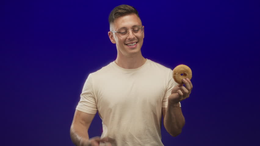 Man wearing glasses and beige t shirt holding glazed donut while smiling and pointing finger to camera in studio; playful joy.