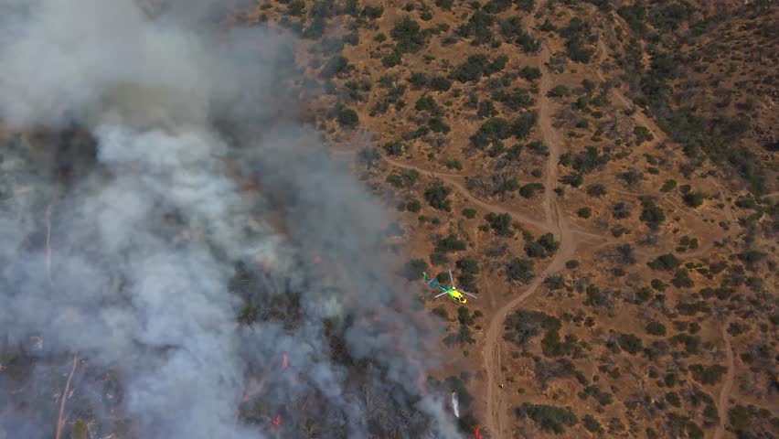 Top-down aerial view of a firefighting helicopter flying over smoldering brush and smoke on a rugged mountain slope in Chile and 4k.