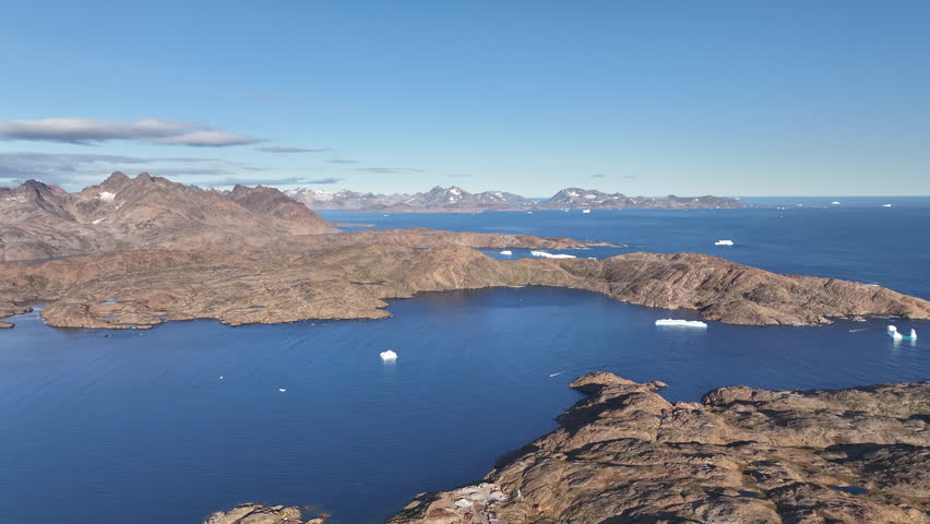 Drone footage looking down at the streets and colorful wooden houses of Tasiilaq, Greenland. The shot reveals the rocky terrain, winding roads, and residential architecture of the remote Arctic settlement during a sunny summer day.