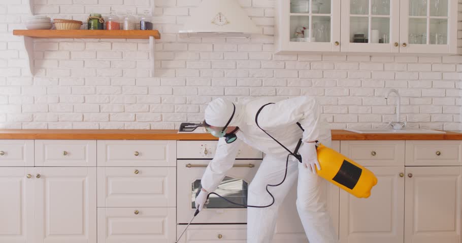 Professional exterminator in white protective suit and mask uses sprayer to treat stove and kitchen cabinets with pesticide for insect control. Pest control services concept