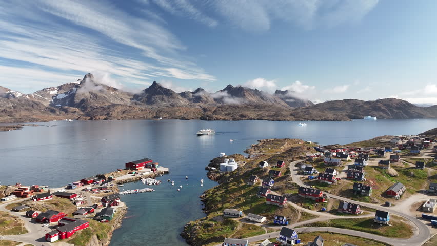 Atmospheric view of Tasiilaq, the largest town in East Greenland. Low clouds hug the jagged mountain peaks overlooking the deep blue fjord, icebergs, and the colorful settlement. Perfect establishing shot for Arctic travel or nature documentaries.