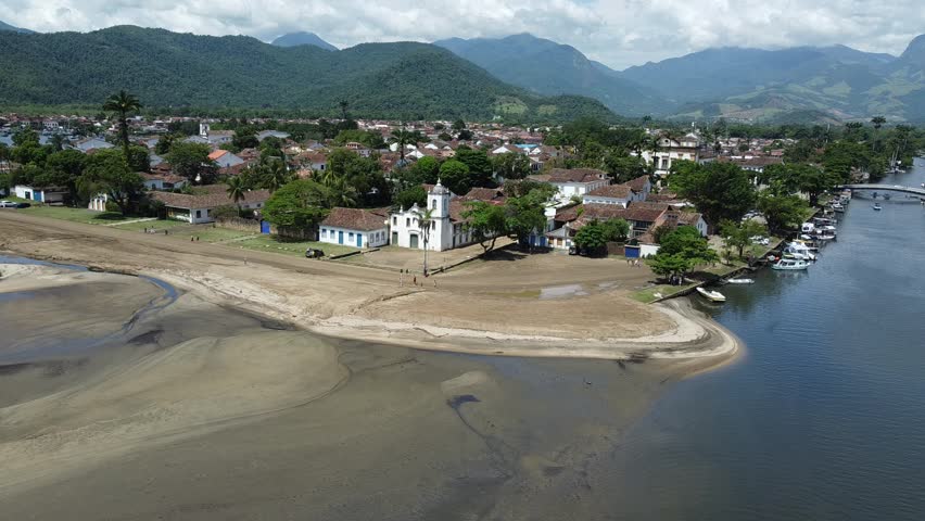 Drone flying from the bay of Paraty towards the town and beach by the river, with the mountains and the Atlantic forest that surrounds this historical city