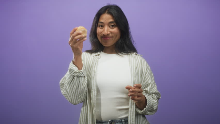 Woman holds muffin and points finger up in studio with purple backdrop, smiling and gesturing toward snack; joyful snack moment.