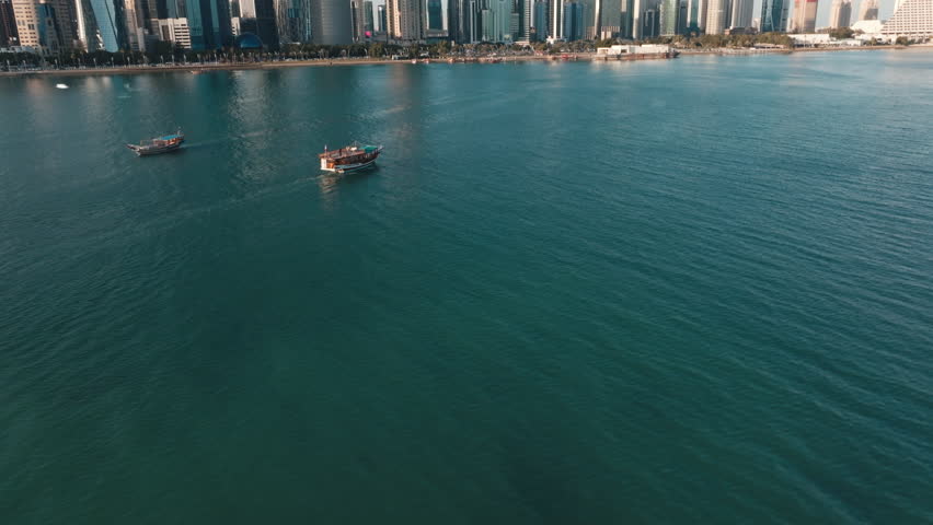 Aerial shot of the Doha skyline with traditional wooden dhows sailing across the turquoise waters of the bay on a sunny day in Qatar