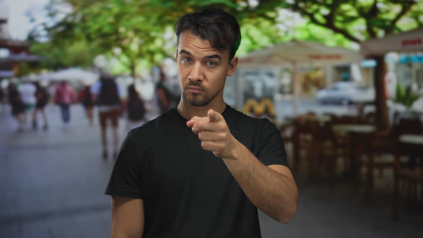Young man outdoors on terrace at restaurant with trees and people in background, playfully pointing and thinking in casual setting
