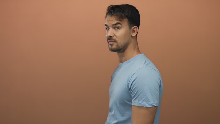 Young hispanic man in blue shirt making excited expressions against brown background, showcasing positive and surprised emotions.