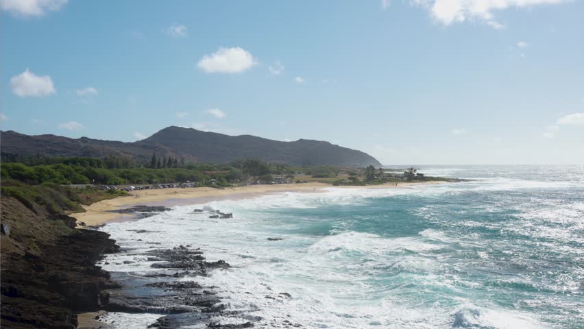 Breathtaking panorama of a tropical island coastline featuring powerful turquoise waves breaking against a rugged volcanic shore and a sandy beach on a beautiful sunny day in hawaii
