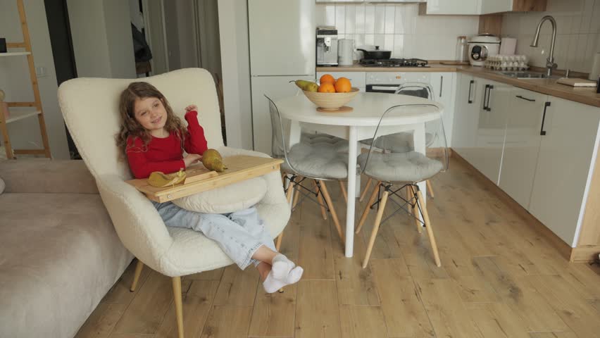A young girl sits in a comfortable chair, enjoying a pear. She smiles and looks happy in her modern home. The bright open floor plan creates a peaceful setting.