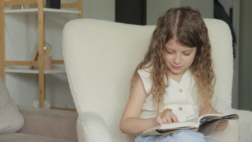 A young girl sits in a fluffy white chair, reading a book and smiling. She looks up and laughs.