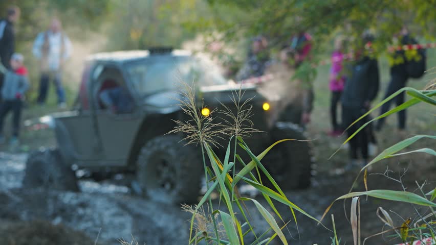 An all-terrain vehicle racing on a muddy off-road track. Selective focus on the reeds in the foreground, while the vehicle and spectators are blurred in the background