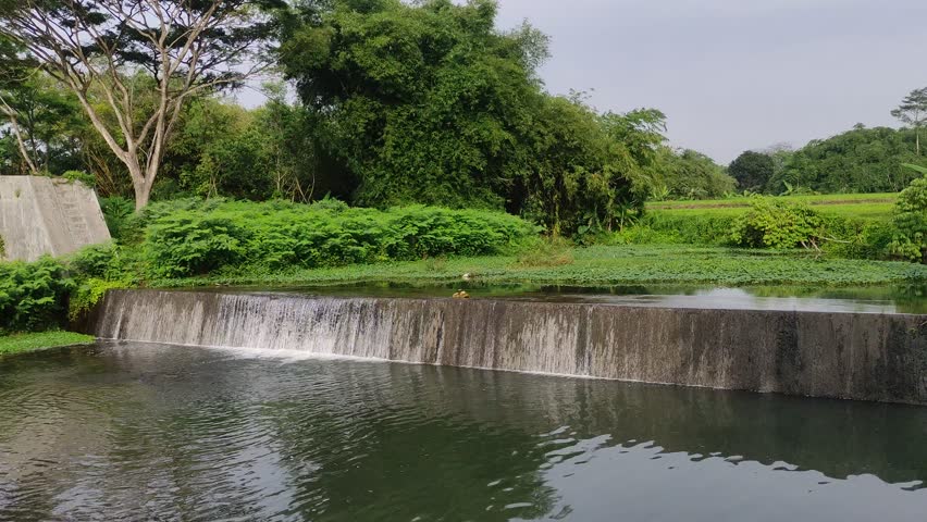 Small concrete dam and waterfall in a rural river with green trees and forest background