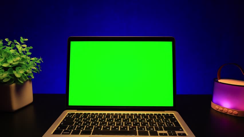 A sleek laptop with a vibrant green screen sits on a black desk next to a potted plant and a colorful speaker, set against a deep blue background