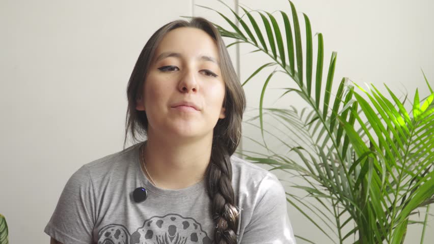 Smiling young woman with a long dark braid, wearing a grey t-shirt and a small lapel microphone, speaking in front of a green palm plant.